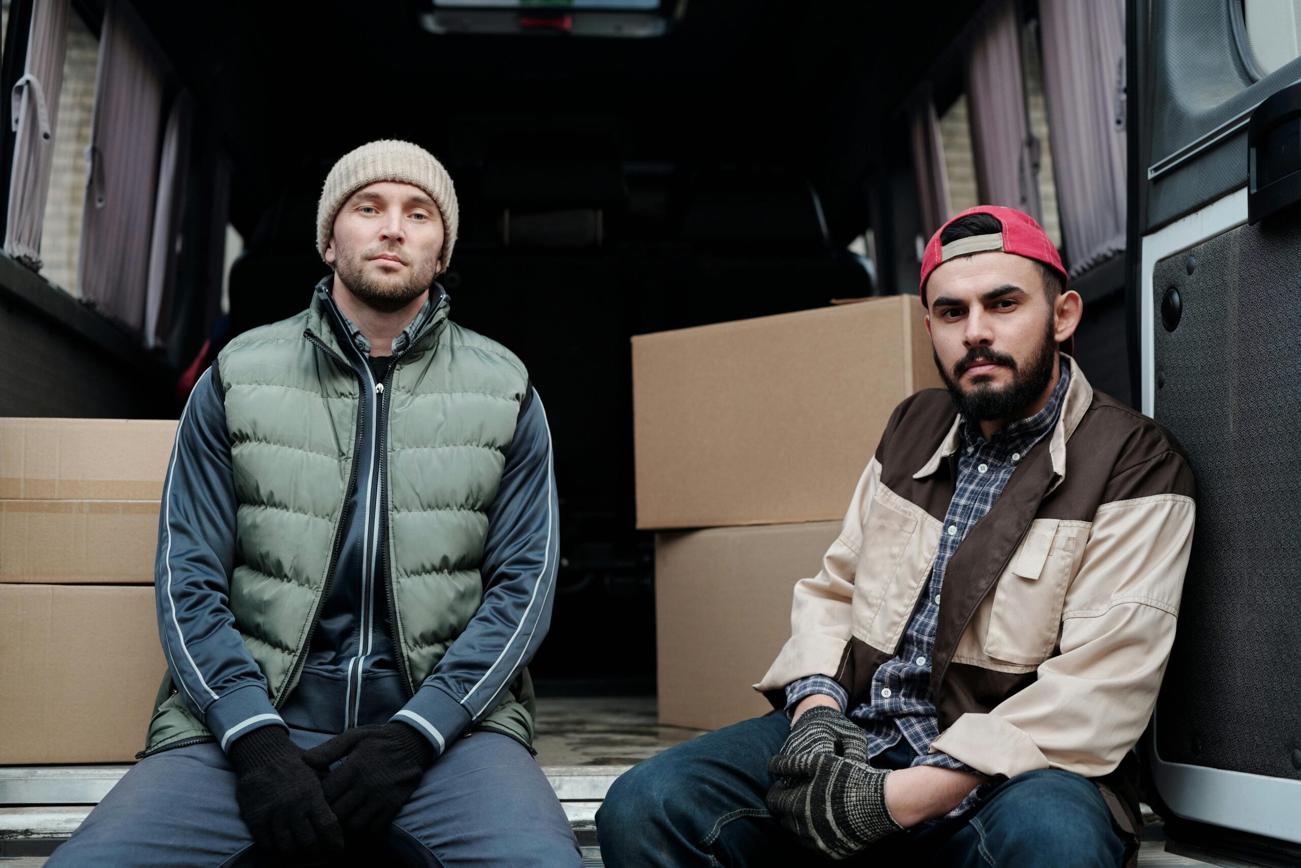 Two men sitting in a van surrounded by boxes, dressed in winter clothing.