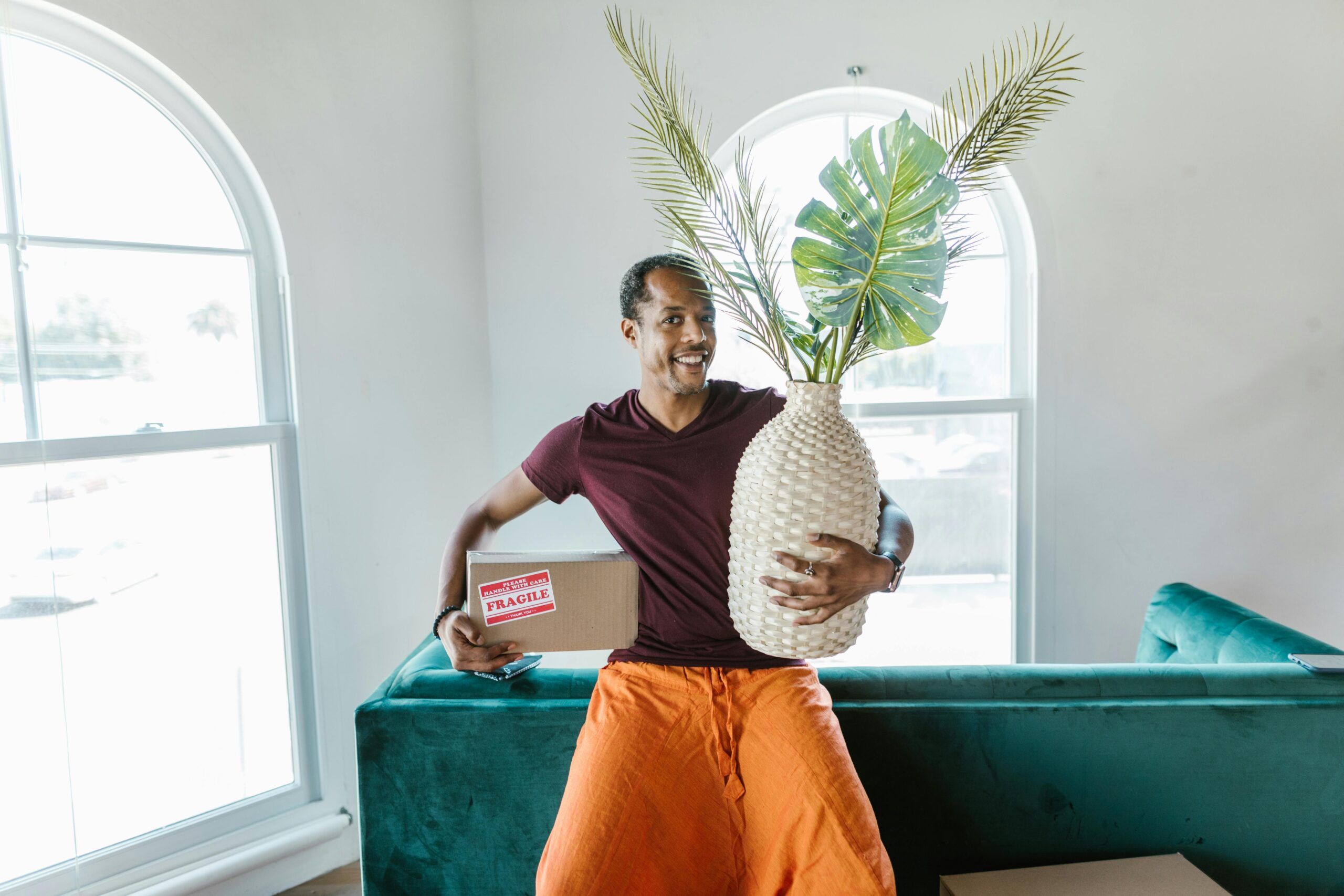 Smiling man holding a fragile box and plant during a home move.